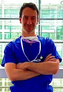 Marco Baia stands in scrubs and a stethoscope with his arms crossed
