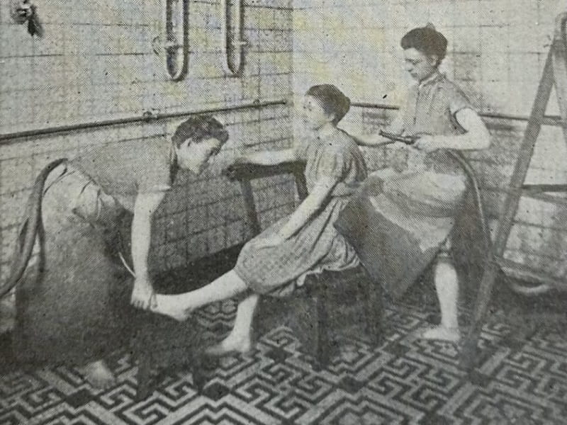 A black and white photograph of a patient sitting on a chair while being massaged and sprayed with water by two attendants. The room is tiled and several pipes are visible.