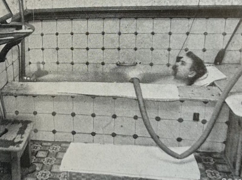 A black and white photograph of a patient in a tiled bath, with a thick pipe running into it.