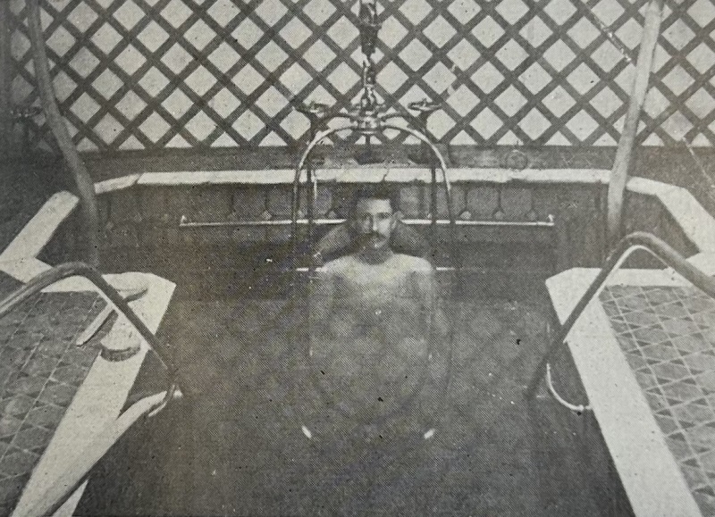 A black and white photograph of a man in a deep, walk-in bath, surrounded by pipes