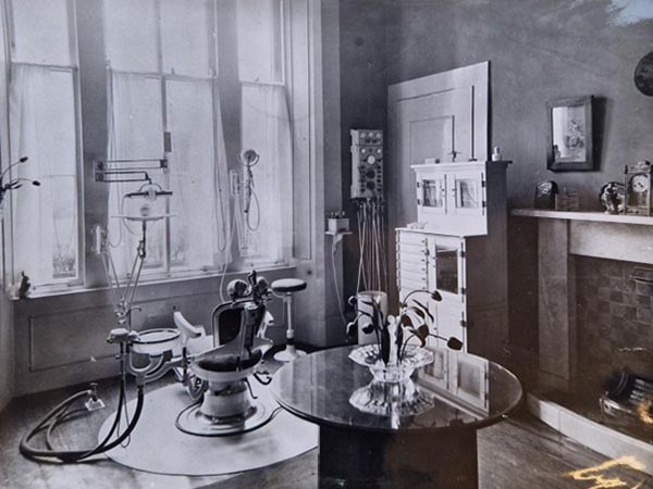 A black-and-white photograph of a dentist's chair in front of a wide window, with associated equipment, a table and flowers, and a fireplace.